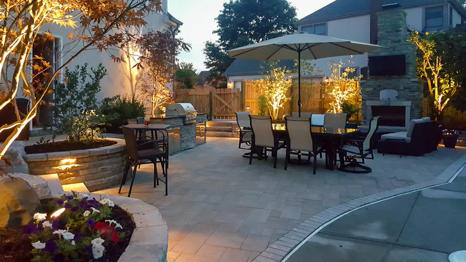 Poolside patio with dining furniture, large planters, tree uplighting, and warm landscape lighting at twilight